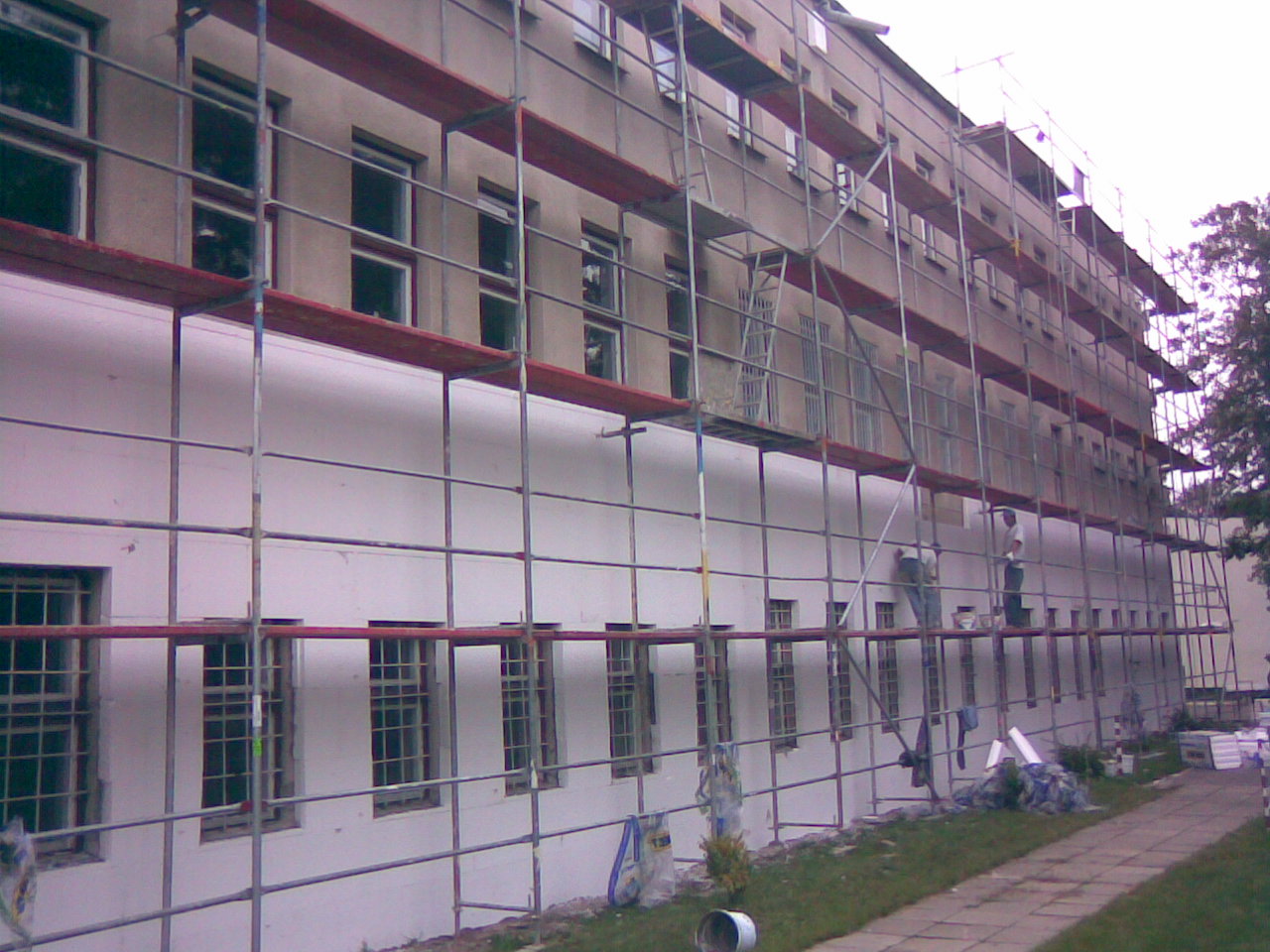 Prace na wysokościach: budynek wielopiętrowy w trakcie docieplania, widoczne rusztowania, pracownicy w białych koszulkach przy pracy, biała elewacja, fragment chodnika i trawnika.